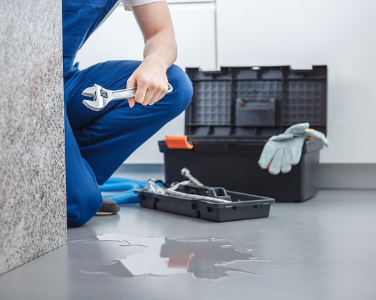 Plombier en salopette bleue avec clé à molette, accroupi devant une fuite d'eau au sol, près de ses outils.