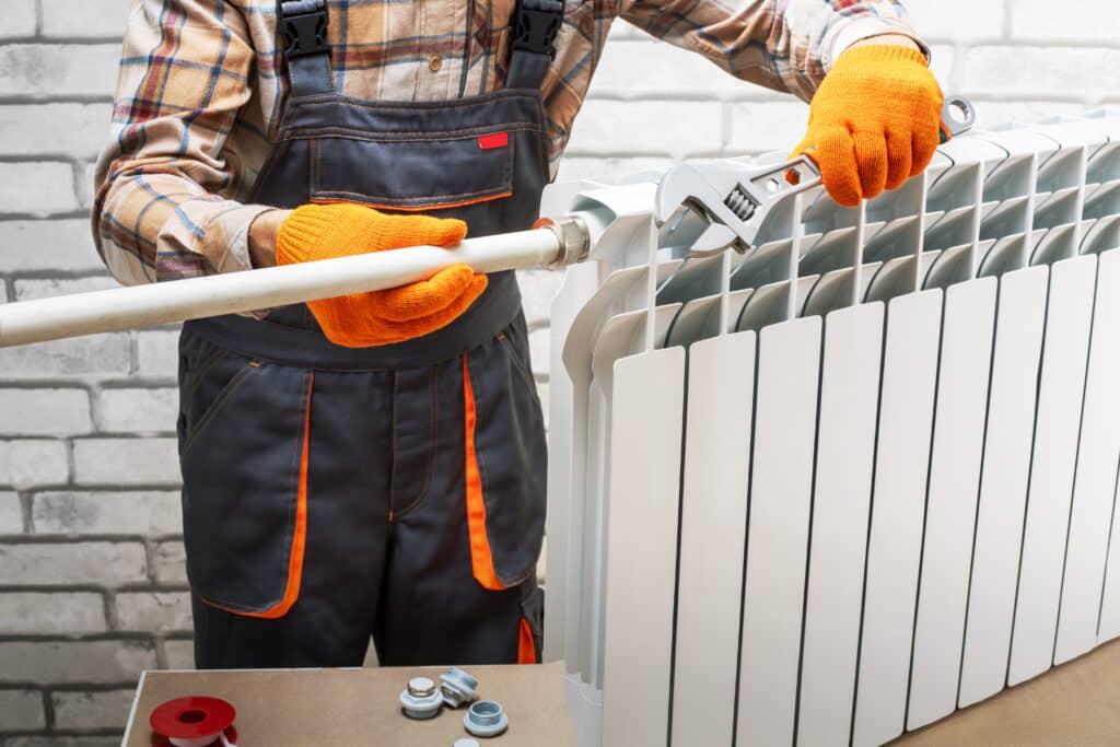 Réparation de Radiateur : Plombier Professionnel en Action Plombier en gants orange réparant un radiateur de chauffage blanc avec une clé à molette. Raccords et ruban PTFE sur un établi.